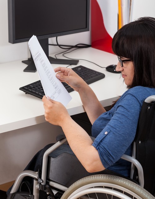 Vrouw in rolstoel met papieren in de hand zit aan haar bureau